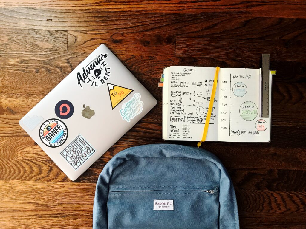 tavolo di legno con zaino blu, computer con adesivi colorati e quaderno con appunti colorati per il ritorno a scuola.